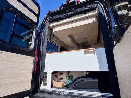 Modern RV rental interior, seen from the rear with doors open. Features clean white/wood design, storage, and ceiling lighting.