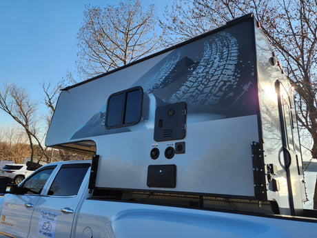 A white pickup truck with a large, graphically designed truck camper, branded as an RV rental unit, parked outdoors.