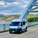 Ram ProMaster RV rental van on a bridge over a scenic lake and mountains under a cloudy sky.