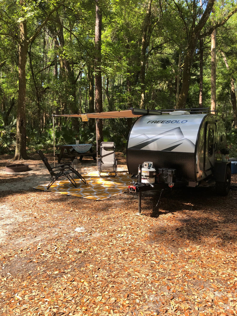 An RV rental camper with an awning, picnic table, and chairs set up in a serene, leafy forest campsite on a sunny day. — photo 13