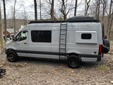Gray Sprinter camper van, ideal for rv rental adventures, featuring a roof rack, ladder, and off-road tires, parked outdoors.