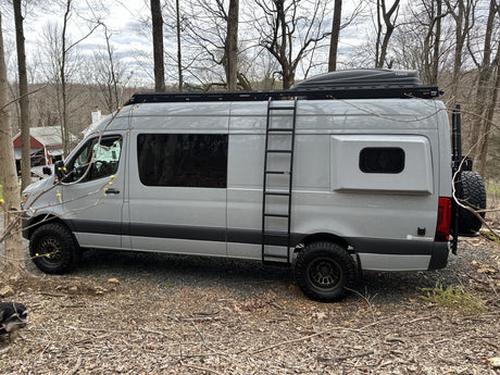 Gray Sprinter camper van, ideal for rv rental adventures, featuring a roof rack, ladder, and off-road tires, parked outdoors.