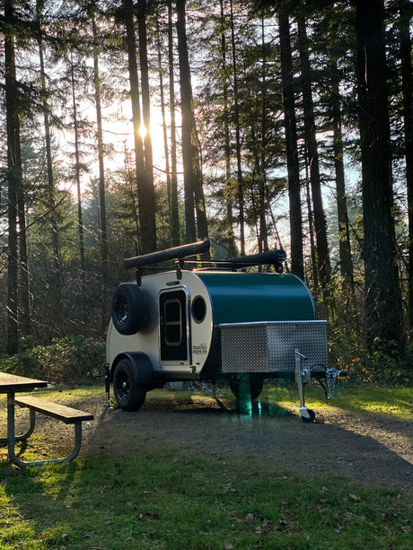 Teardrop camper trailer, ideal for an RV rental, parked in a sunlit forest campsite with a picnic table.