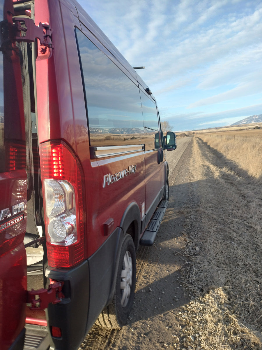 Scenic rv rental: Red Pleasure-Way camper van on a dirt road. Rear doors open, mountains reflected in side windows.