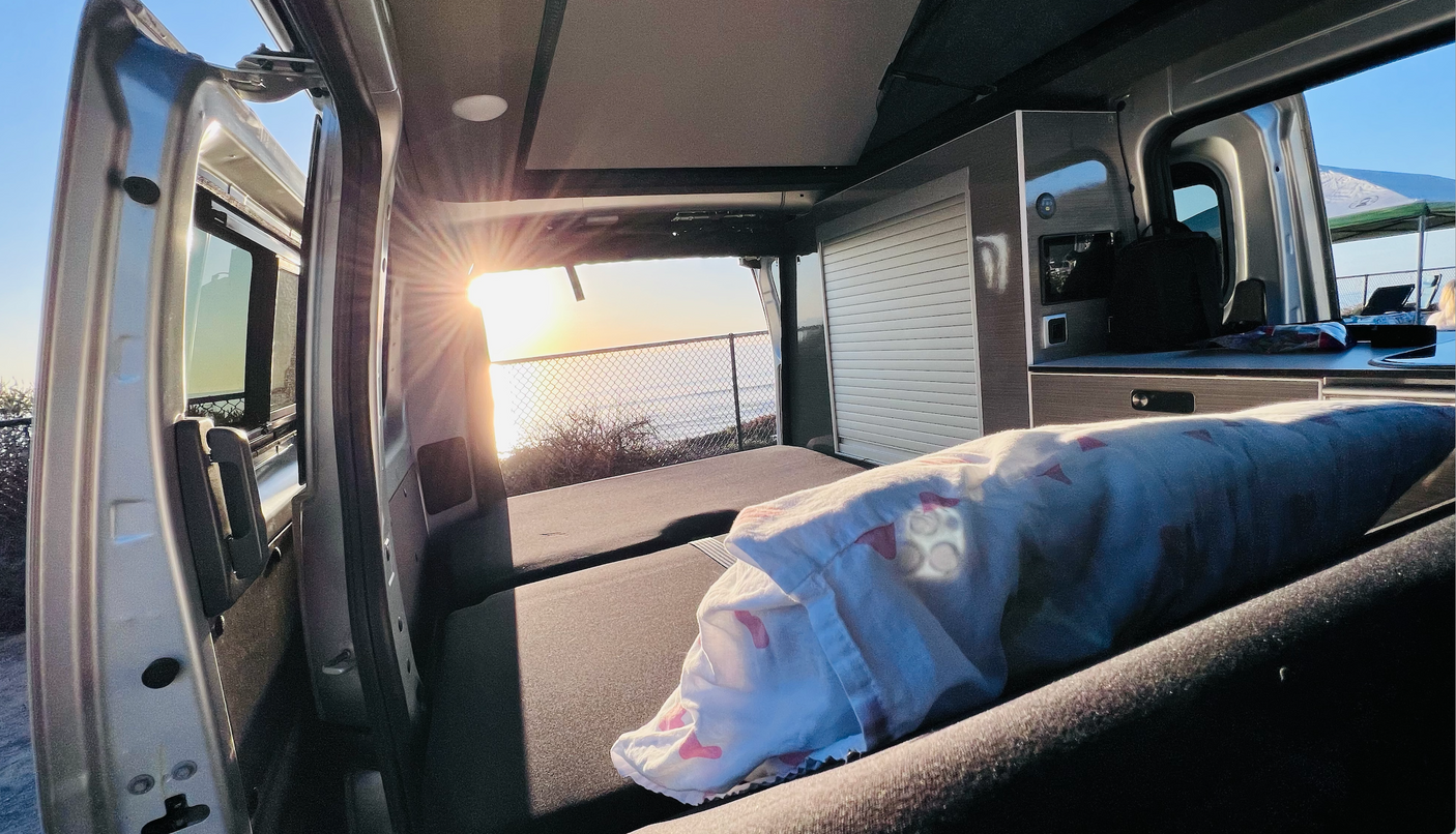 Bright rv rental interior at sunset. A bed and kitchen unit face open doors, revealing an ocean view and setting sun.