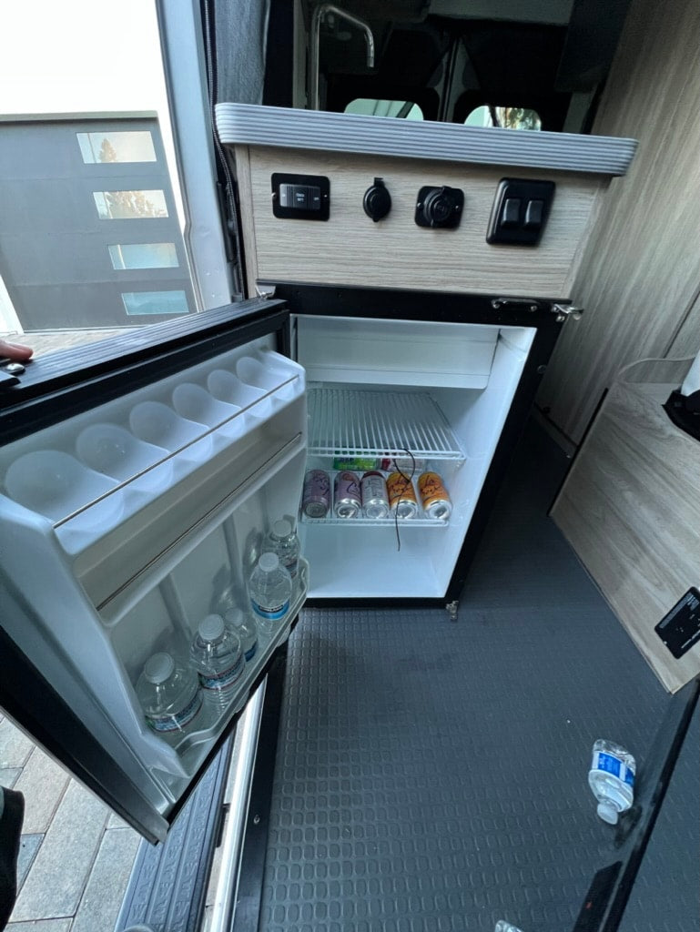 RV rental kitchen: open mini-fridge with drinks/water, showing power outlets & controls on the cabinet above. — photo 13