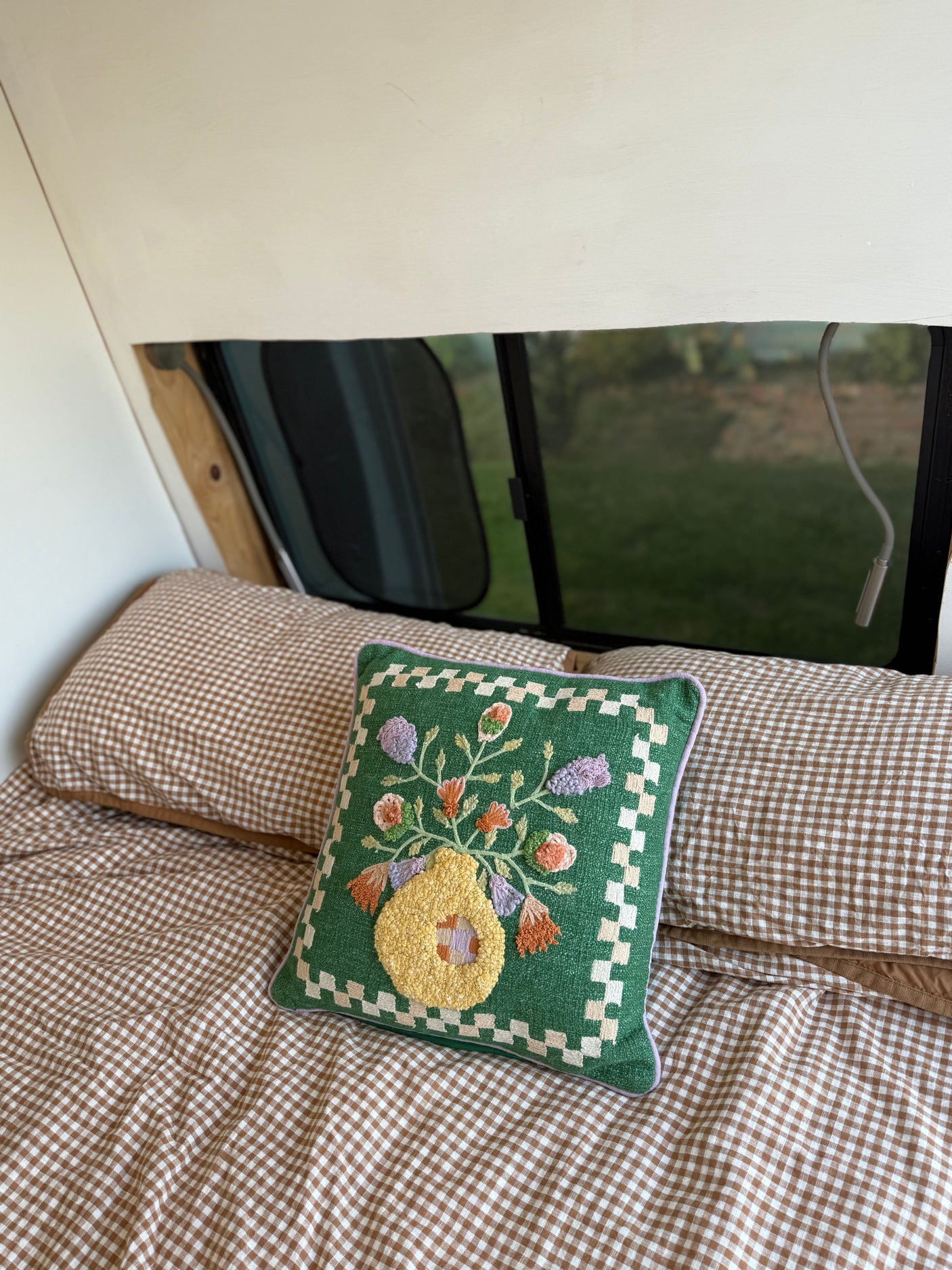 Cozy RV rental interior with brown gingham bedding and a green, floral embroidered pillow by the window.