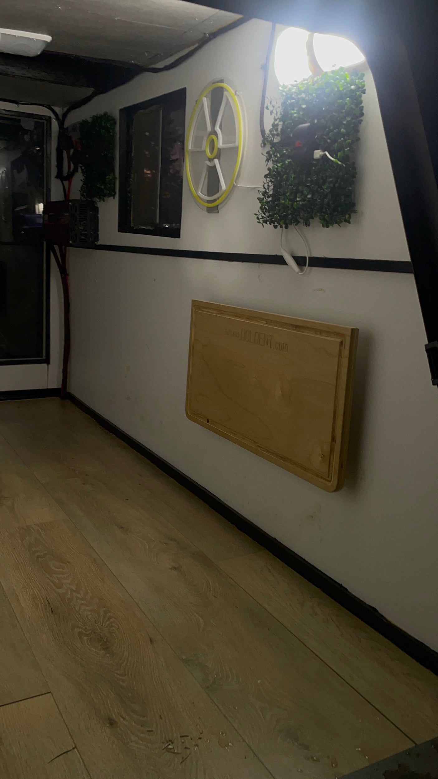 An RV rental's cozy interior features light wood flooring, white walls with a mounted wooden folding table, neon light, and plants.
