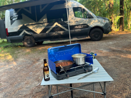 RV rental: Camp cooking a meal & boiling water on a portable stove, with beer, at a forest campsite by the camper van.