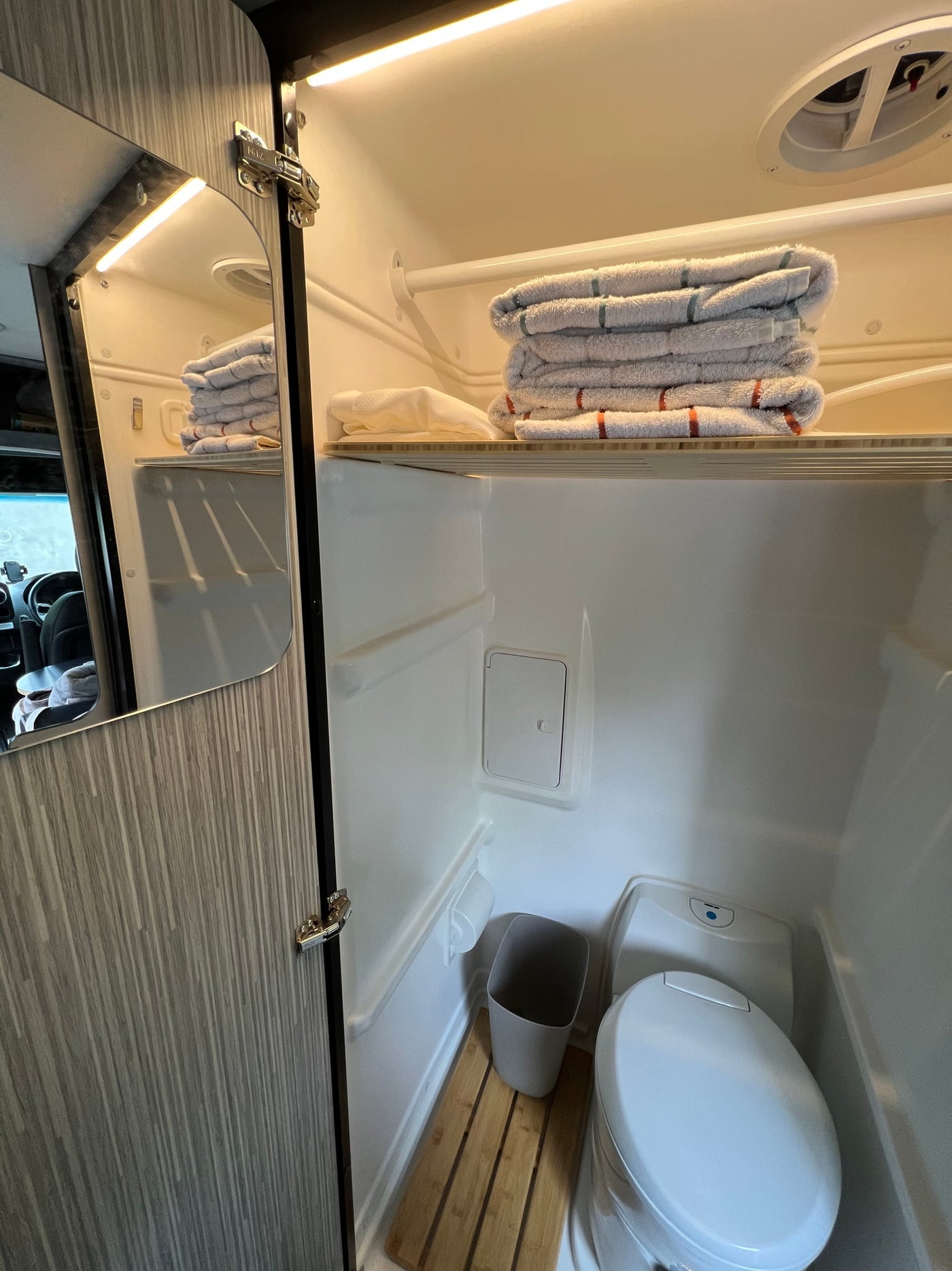 Inside an RV rental's compact bathroom: toilet, trash can, wooden mat, and neatly folded towels on a shelf. — photo 13
