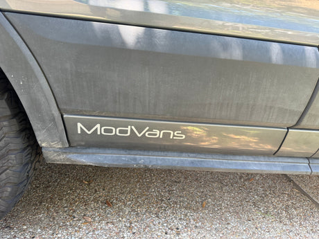 Dusty gray ModVans RV rental exterior, featuring the custom logo on the side body panel near the tire.