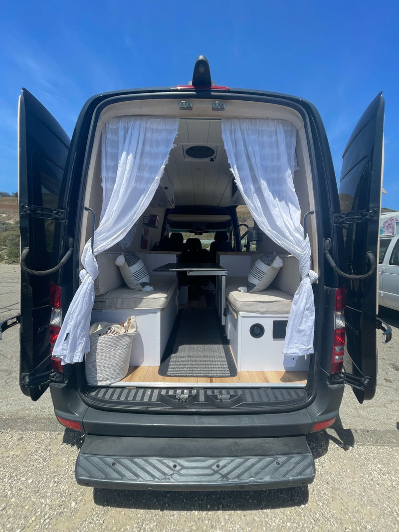 Open back of a black RV rental, revealing a cozy camper van interior with bench seats, table, and white curtains. — photo 7