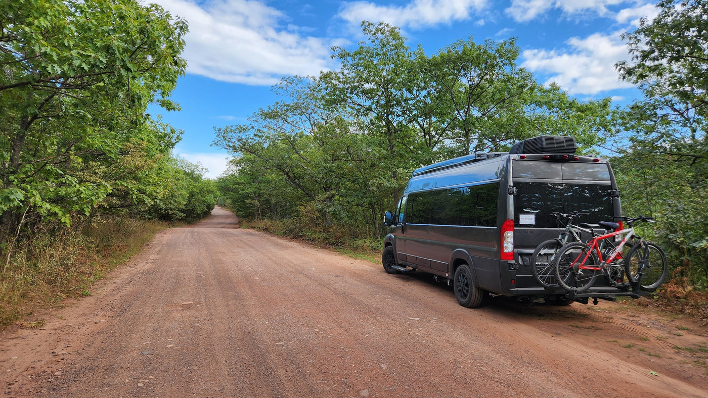 Adventure awaits! Dark grey RV rental parked on a dirt road in a sunny forest with bicycles on the back, ready for exploration. — photo 10