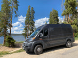 Dark gray rv rental camper van parked by a serene lake amid pine trees under a blue sky with white clouds.