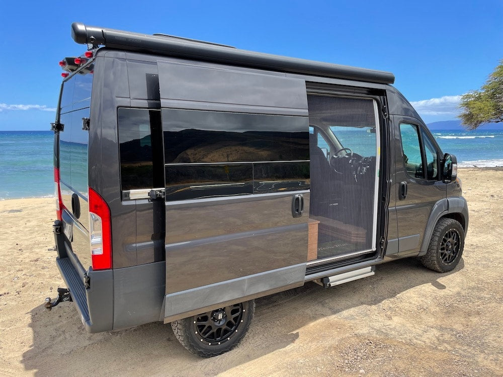  parked on grass in a lush setting. Perfect for an RV rental adventure!|Dark grey camper van RV rental parked on a beach with its side door open — photo 5
