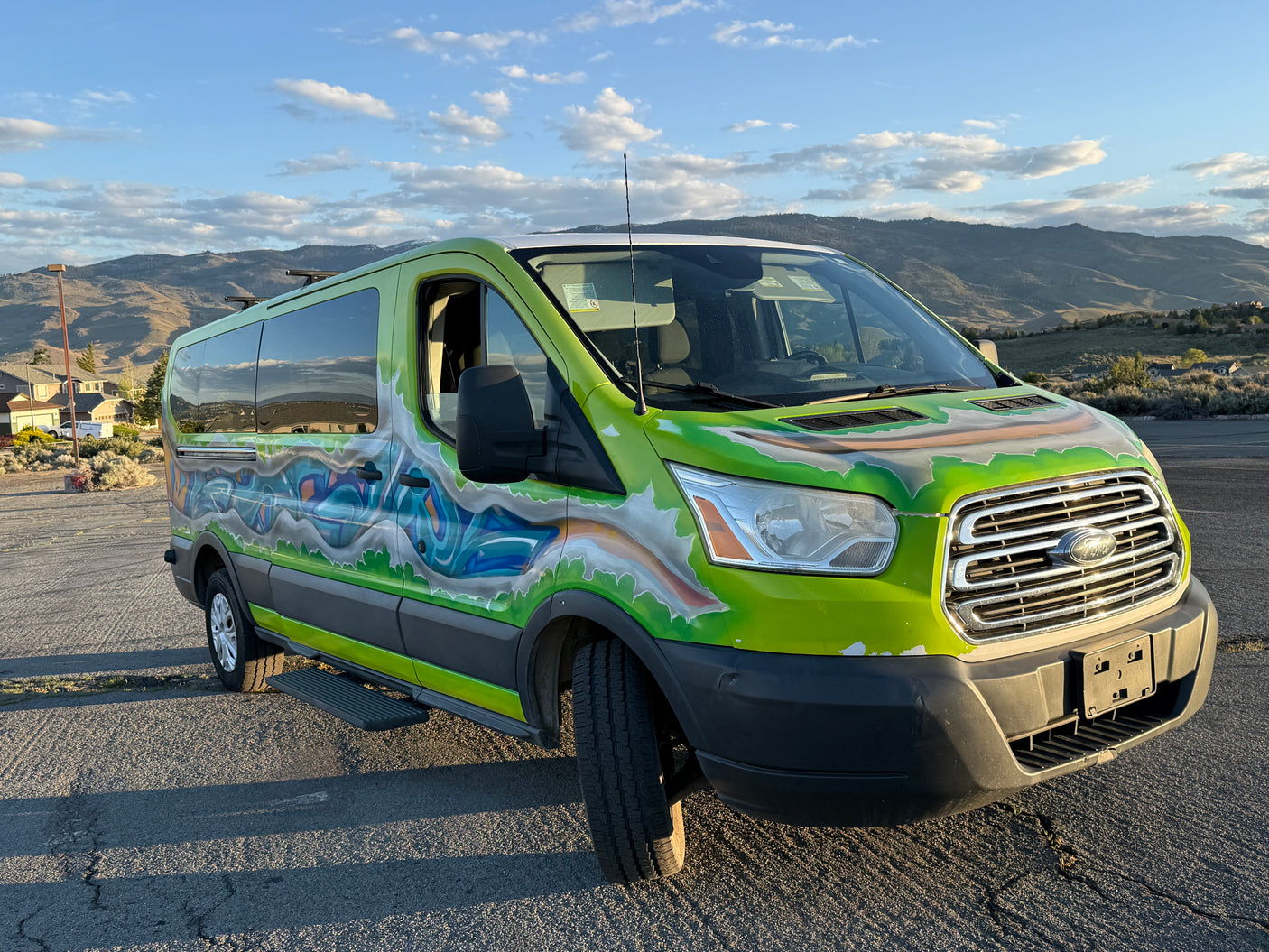 Bright green Ford Transit RV rental, custom-painted with graffiti art, parked outdoors with mountains & blue sky.
