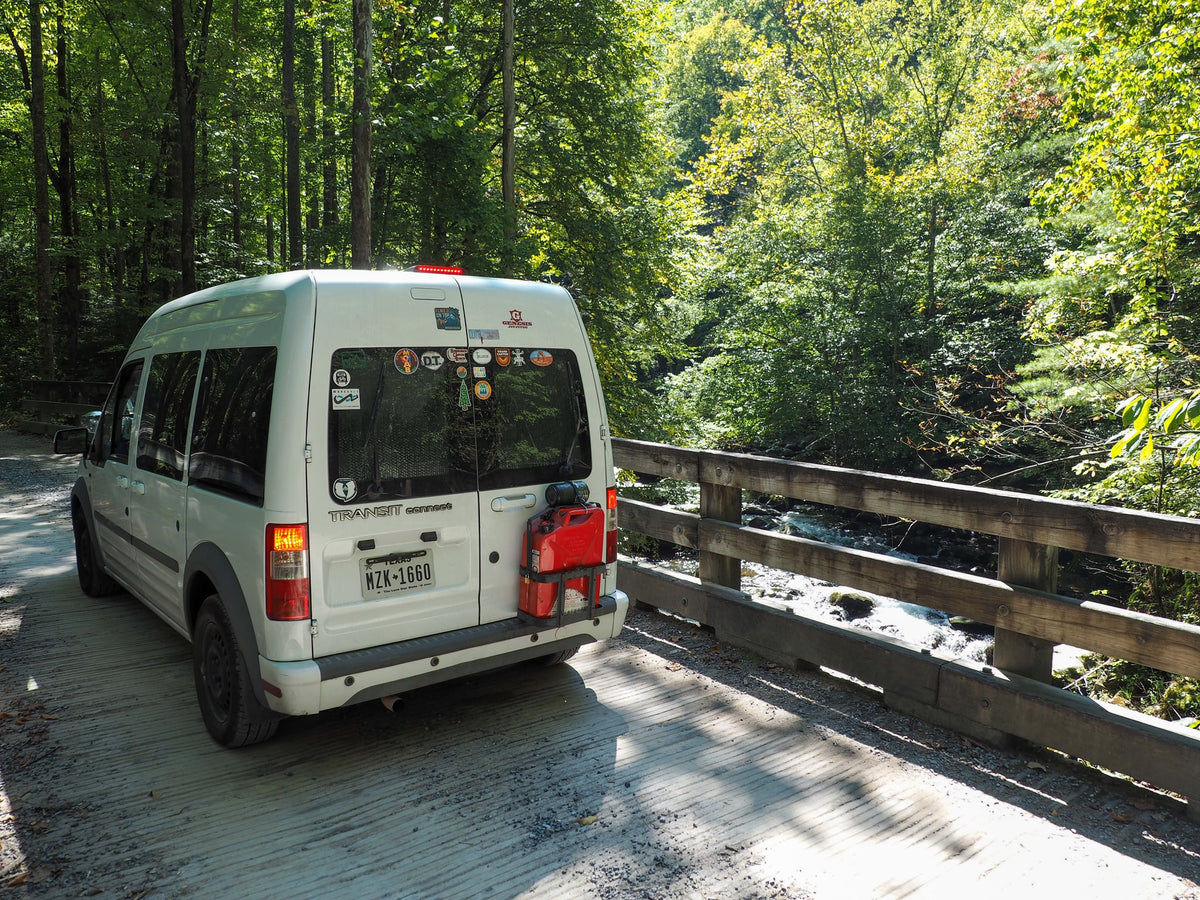 RV rental: Ford Transit Connect campervan on a scenic bridge over a stream in a lush forest, ideal for van life.