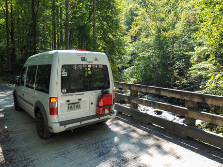 RV rental: Ford Transit Connect campervan on a scenic bridge over a stream in a lush forest, ideal for van life.