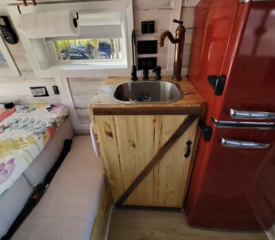 Inside an RV rental: a compact kitchen with a wooden counter, sink, and red retro fridge, next to a comfy bed.