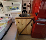 Inside an RV rental: a compact kitchen with a wooden counter, sink, and red retro fridge, next to a comfy bed.