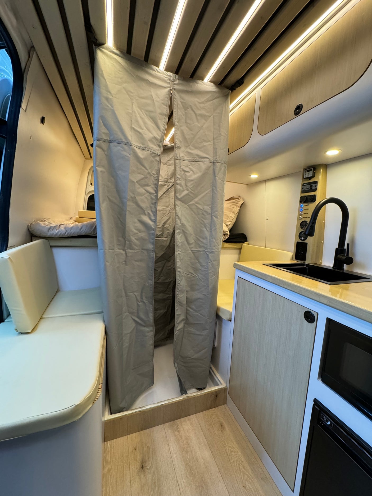 Compact RV rental interior featuring a convertible bed, kitchen with sink, microwave, and a private shower area.