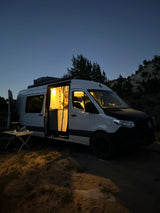 A cozy RV rental camper van glows at night, parked in a natural setting. Person visible inside, outdoor table ready.