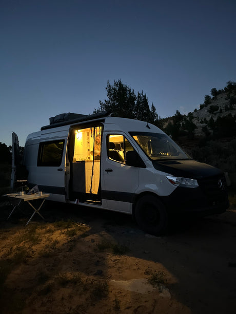 A cozy RV rental camper van glows at night, parked in a natural setting. Person visible inside, outdoor table ready.