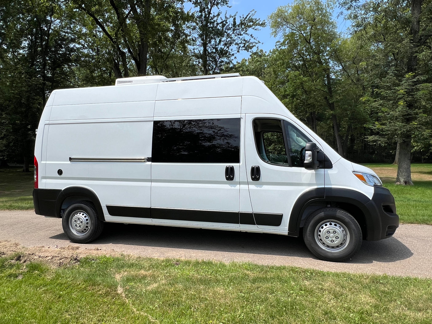 White high-roof Ram ProMaster camper van for RV rental, parked on a sunny path with trees.