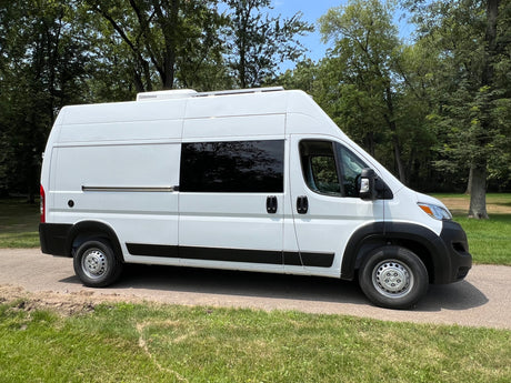 White high-roof Ram ProMaster camper van for RV rental, parked on a sunny path with trees.