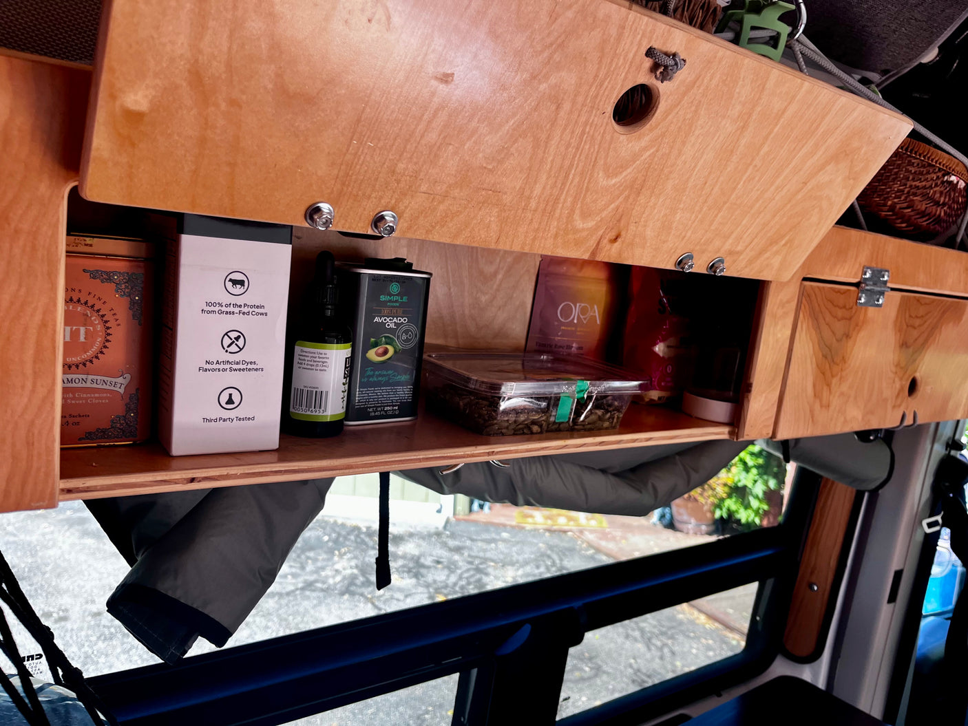 RV rental interior: Open wooden cabinet in a van, showing stored tea, avocado oil, and health supplements for travel convenience.