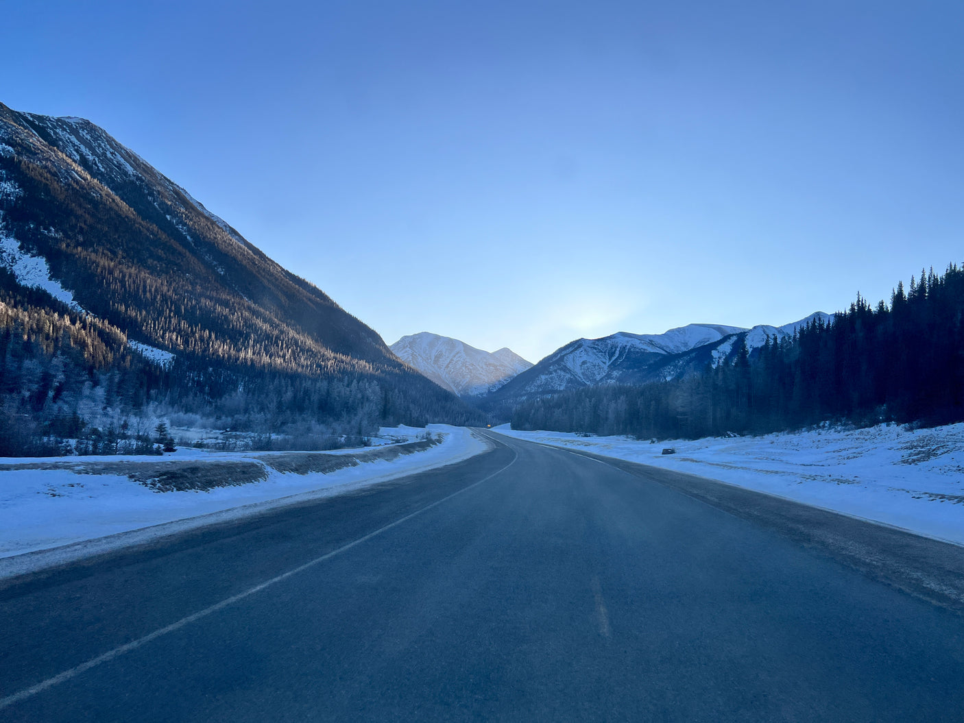Experience a snowy mountain **rv rental** road trip! Clear highway winds through a valley with forested peaks under a bright blue sky.