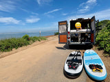 RV rental by tranquil waters: A white camper van with open doors and a person inside, plus two paddleboards ready for adventure.
