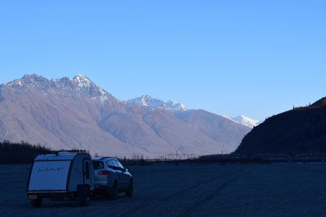 Explore scenic mountains with an RV rental! A teardrop trailer & SUV are parked on a gravel bar by bridges.