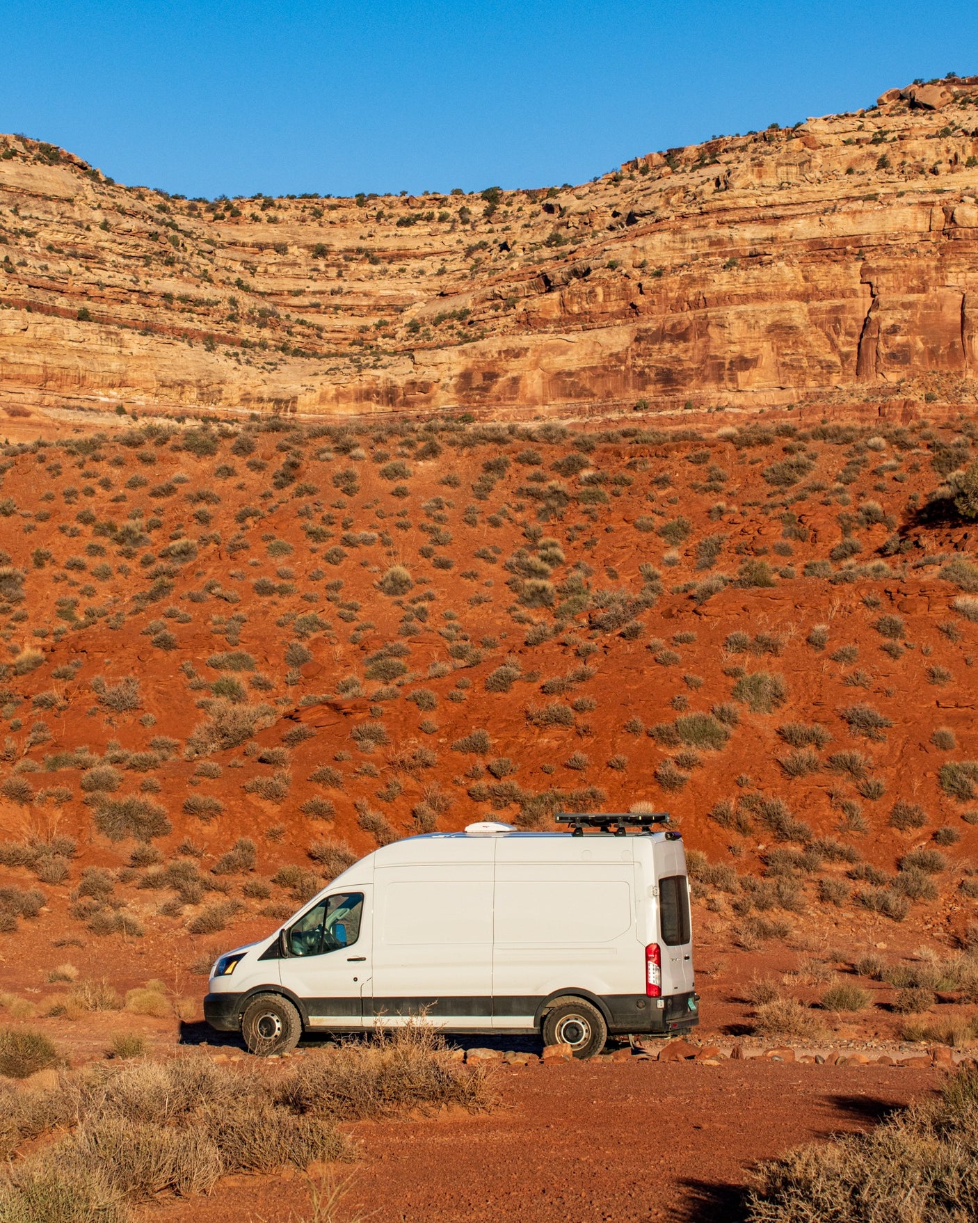 White rv rental camper van parked in a stunning red desert with layered rock formations under a clear blue sky.