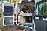 Rear view of an RV rental: a man relaxes inside on a bed, protected by a mesh screen, parked in woods.