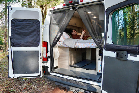 Rear view of an RV rental: a man relaxes inside on a bed, protected by a mesh screen, parked in woods.