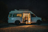 Starry night van life: Woman enjoys cozy RV rental interior under a brilliant, star-filled sky. Illuminated gravel road.