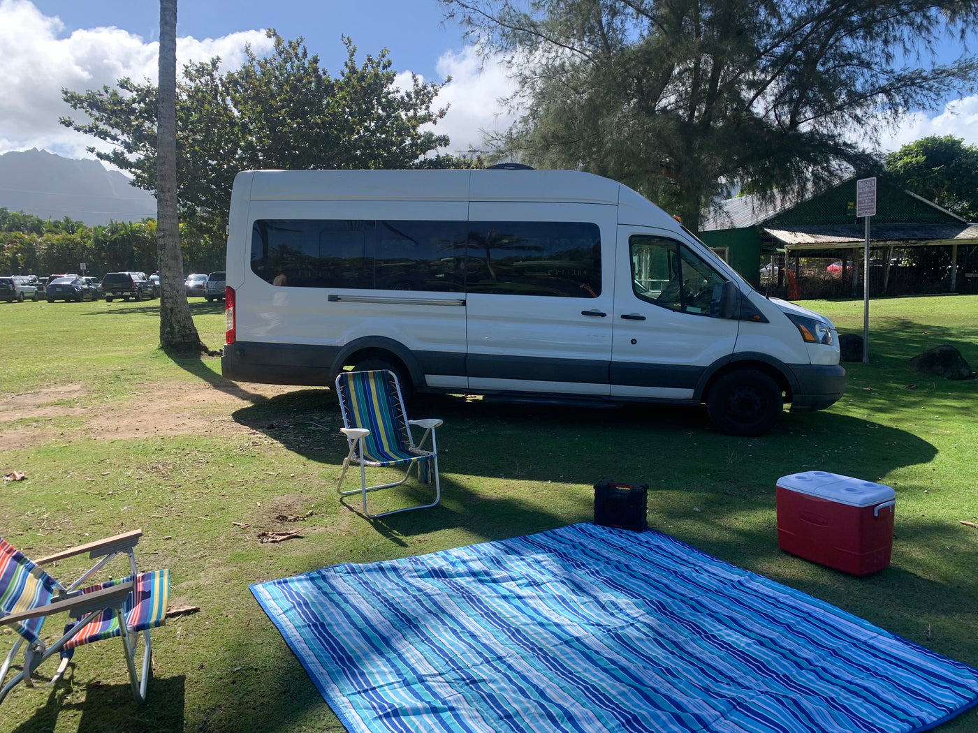 RV rental van parked on green grass, set up for a picnic with a striped blanket, chairs, a cooler, and a speaker.