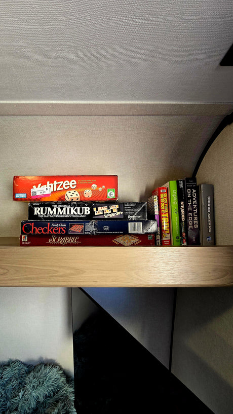 A cozy rv rental shelf is stocked with classic board games like Yahtzee, Rummikub, Scrabble, and various books for entertainment.
