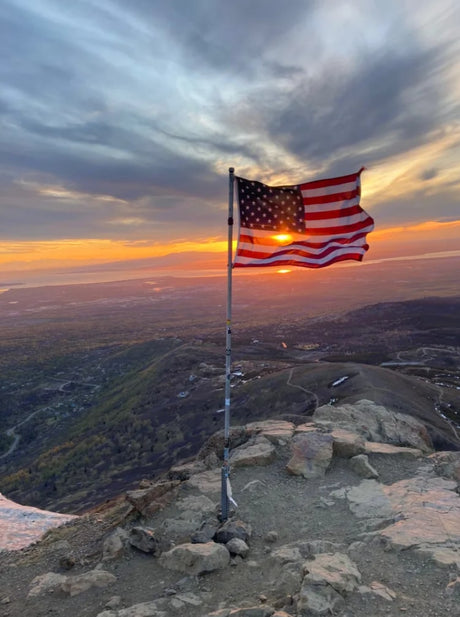 American flag waves at a mountain summit, sun-kissed at sunset, overlooking a vast valley. Your RV rental adventure!