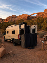 Camper van RV rental with outdoor shower setup at a desert campsite, backlit by a stunning natural arch.