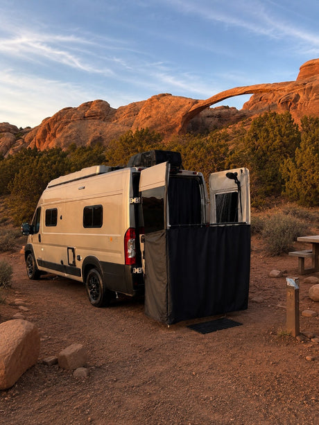 Camper van RV rental with outdoor shower setup at a desert campsite, backlit by a stunning natural arch.
