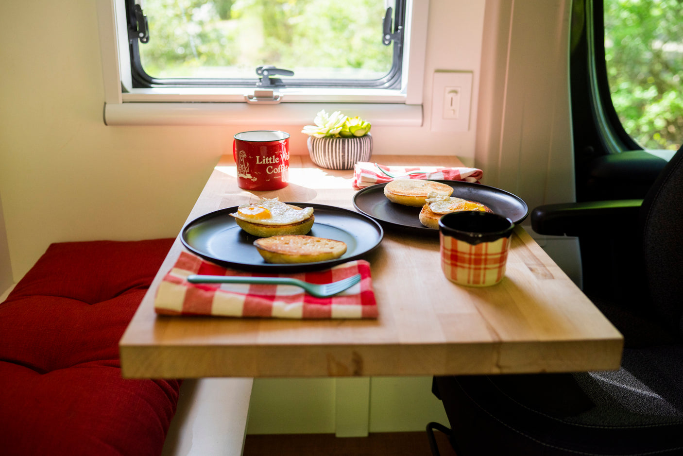 An RV rental's cozy table with two plates of breakfast: English muffins, eggs, and coffee. Bright window view.