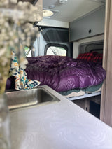 Inside a compact RV rental: a plush purple bed, kitchen sink, and functional overhead cabinets.