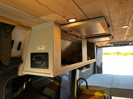 Inside a custom RV rental van: wooden ceiling, overhead cabinets with dishes, a solar controller (13.5V), sink, & bed.