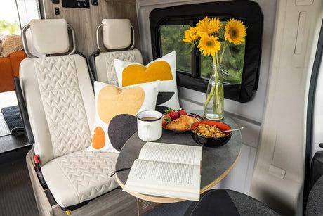 Enjoy a delightful breakfast inside this cozy RV rental! Comfy seats, a table with food, book, and sunflowers by the window.