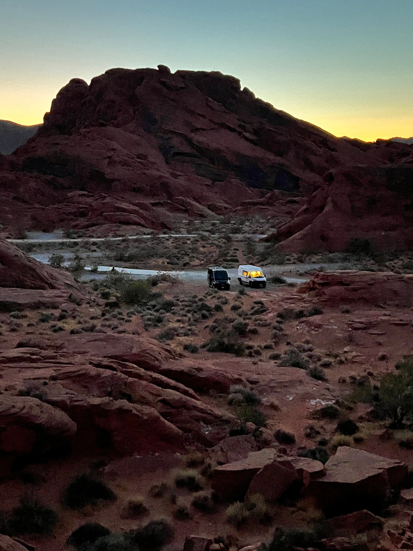 Sunset over red rock mountains, with an RV rental glowing inside, and another van parked for desert camping.