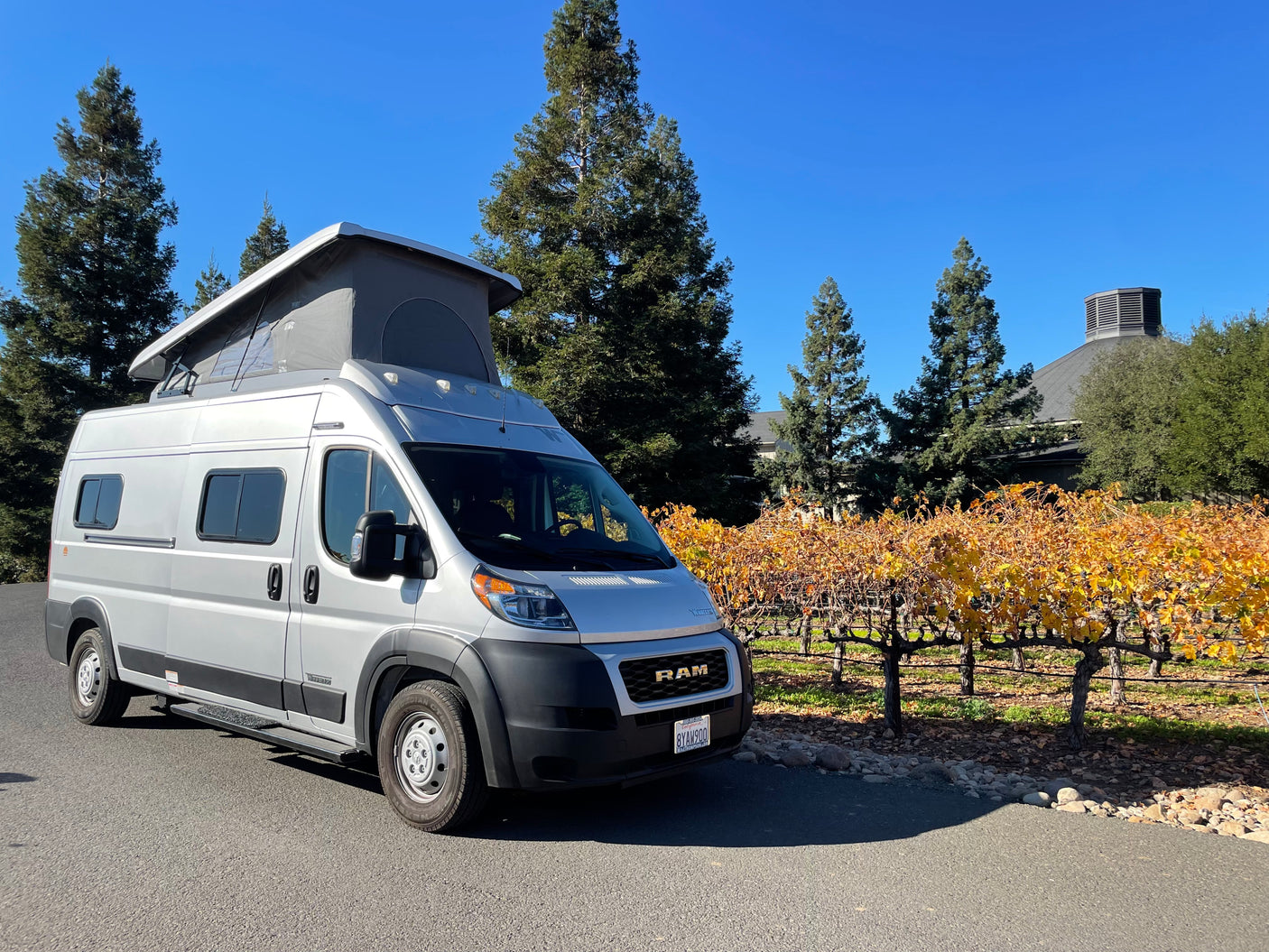 Silver RV rental, a pop-top camper van, parked beside a vineyard with vibrant fall foliage and tall trees on a sunny day.