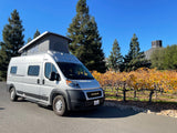 Silver RV rental, a pop-top camper van, parked beside a vineyard with vibrant fall foliage and tall trees on a sunny day.