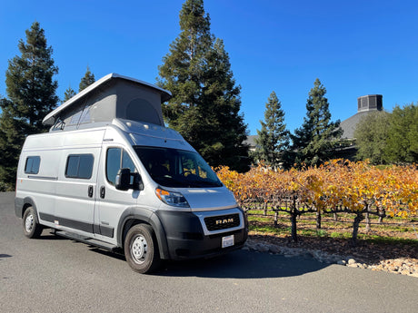 Silver RV rental, a pop-top camper van, parked beside a vineyard with vibrant fall foliage and tall trees on a sunny day.